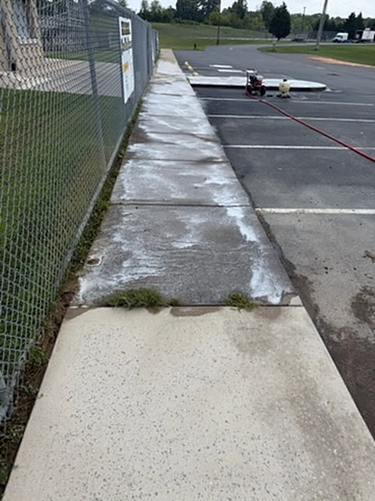 Concrete walkway beside a ball field fence before washing, darkened with grime and weeds coming through the joints